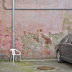 Backyard of old weathered grunge house. Pink cracked brick wall, white chair and black car