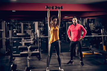 Slim woman doing exercises with dumbbells while personal trainer watching her. In background exercise equipment.
