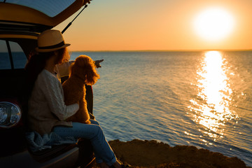 Woman traveler with dog sitting in car trunk near sea, watching sunset.