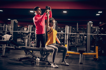 Woman sitting on a bench while personal trainer helping her to lift bumbbell. Gym interior.