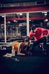 Fotobehang Woman doing planks and her personal trainer kneeling next to her. Side light, gym interior. © dusanpetkovic1