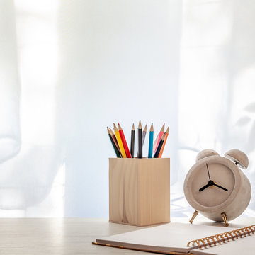 Photo of a white desk with colored pencils in a wooden toolbox, open notebook and a concrete alarm clock on the background of sun-lit curtains. Copy space
