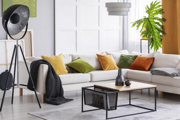 Lamp next to white corner settee with cushions in bright apartment interior with table. Real photo