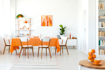 White chairs at table with orange cloth in modern dining room interior with plant and poster. Real photo
