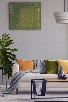 Lamp above wooden table in grey flat interior with green painting and plant next to sofa. Real photo