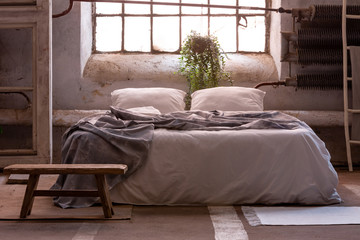 Real photo of a wabi sabi bedroom interior with a bed, plant and wooden stool in front