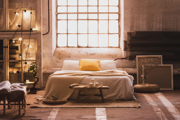 Real photo of a cozy, industrial bedroom interior with a double bed, yellow pillow, window and chain lights - obrazy, fototapety, plakaty