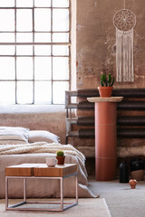 Table in front of a bed, stand with a plant and iron radiator in an industrial bedroom interior. Real photo