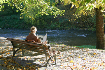 Woman sitting and reading on a bench in the autumn reads a newspaper while relaxing in the sun