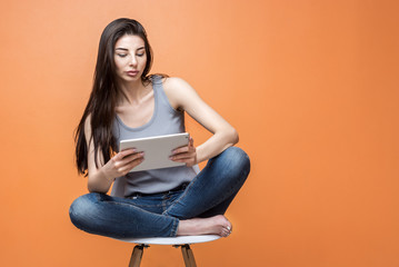 Obraz na płótnie Canvas Portrait of a young beautiful girl with a tablet in her hands sitting in the chair against orange background. Lifestyle, people and technology concept