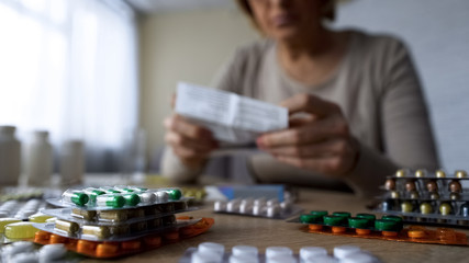 Medication lying on table, sick old woman reading prescription behind, closeup