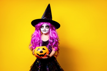 Small girl in Halloween witch dress and hat scaring and making faces isolated on yellow background. Trick or treat.