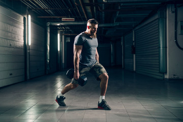 Side view of muscular attractive caucasian bearded man doing lunges with dumbbells in underground...