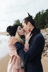 Young pretty sensual couple hugging and kissing at the beach. Seaside coastline and forest on the background. Cold autumn weather.