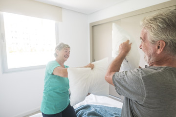 Happy caucasian aged senior adult couple enjoy the early morning making a pillows war together playing and laughing a lot. love and married forever life concept with joyful and playful  mature people