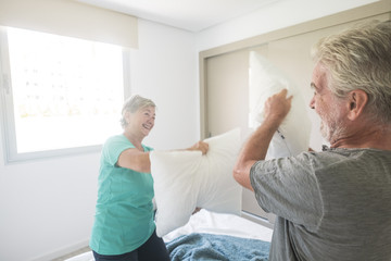 Happy caucasian aged senior adult couple enjoy the early morning making a pillows war together...