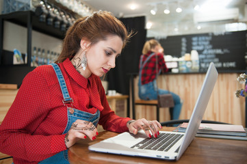 cute young caucasian female sitting in coffee bar pressing keyboard of silver laptop