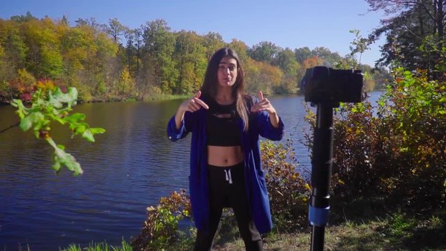 Blogging young woman stand next to camera at nature talking and gesturing slow motion showing thumbs up and smiling
