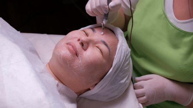 Galvanic cleaning of the skin. Asian woman on the procedure of disincrustation. The work of a beautician with an electrical apparatus in hands. Oriental girl at the reception in a medical beauty salon