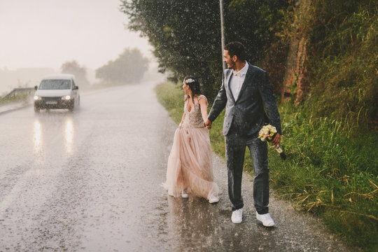 Totally wet just married couple walking on rain by drive road. Watching cars passing by.