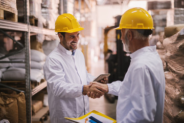 Business is done. Two satisfied smiling males shaking hands after closing a successful business deal. Standing in big warehouse dressed in white coats with helmets on their heads.