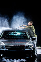 A man with a beard or car washer washes a gray car with a high-pressure washer at night in on the...