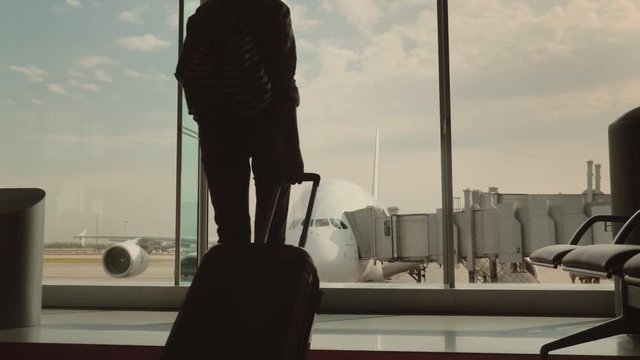 A woman with luggage goes to a large window in the airport terminal. Outside the window you can see a beautiful airliner. In anticipation of the journey