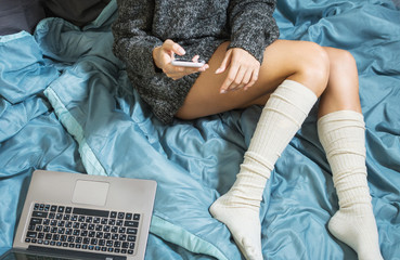 A young girl with sexy legs in a warm sweater lying on the bed with smartphone and laptop. Work at home