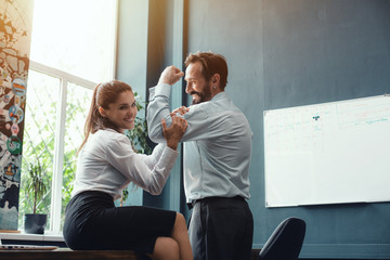 Young woman sitting on desk in office and touching muscles of her male colleague. Fight of genders...