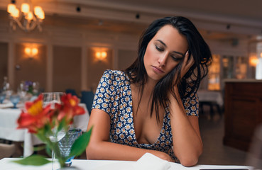 Portrait of tired brunette young elegant woman wearing dress sitting in the restaurant looking down waiting her lunch. Caucasian female posing in cafeteria and waiting her friends. Copy space