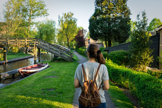 Jovem de costas ao pé de um canal com um barco vermelho e uma ponte de madeira