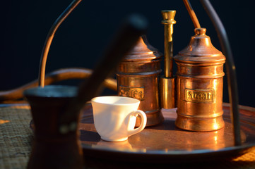 a Cup of coffee near the sugar bowl and a container with coffee on a copper tray and a rattan table