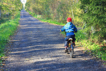 Unidentified Young Boy Riding Bike Alone Outdoors on Rural Road. Child Biking on Empty Long Path, Back View Image. Late Summer Scene with Teen Boy on the Bicycle Moving Forward