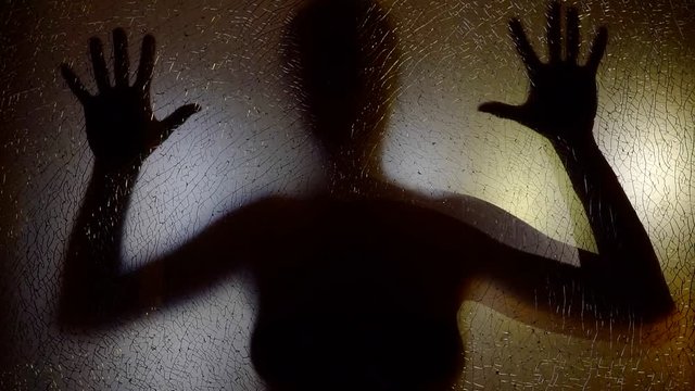 scary movie shot of woman is knocking y both hands with open palms in glass screen in darkness, silhouette view