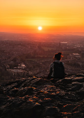 The girl from the back is sitting on a rock of a high mountain watching the sunset over the cit