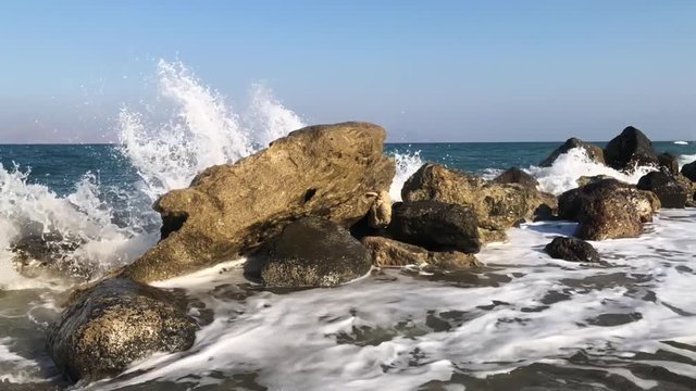 Slow motion shot of ocean waves hitting rocks in the water and exploding in particles towards the camera.
