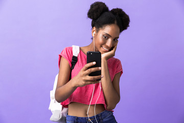 Photo of positive african american woman student wearing backpack and white headphones holding smartphone, isolated over violet background
