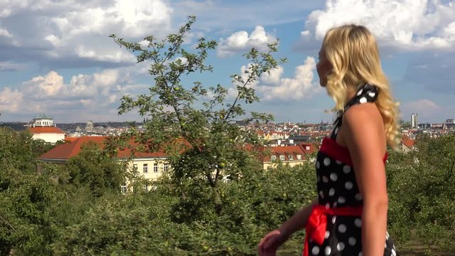 A young beautiful woman in a retro dress down a pathway on a hill and looks at the cityscape in the background