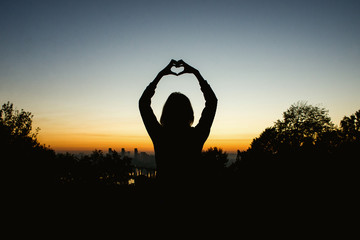 Silhouette of a young girl with hearts made of fingers against the blue sky with clouds at sunset. Lovers in nature.Lovers silhouette. Silhouette of the heart. People and lifestyle concept.