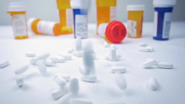 White narcotic pills fall in slow motion with prescription pill bottles in the background. The painkillers represent the opioid crisis in America, addiction, overdoses, and the pharmaceutical industry