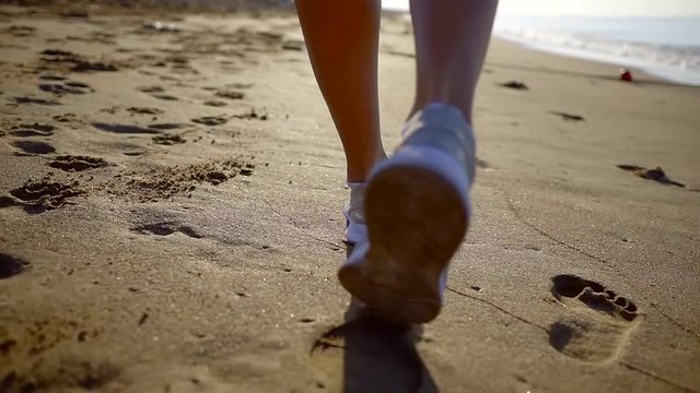 woman is walking on wet sand near sea water in sunny day, leaving traces of her sporty shoes