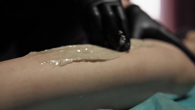 A sugaring is performed using the sugar paste from the legs of a young woman at a spa. The master in black gloves removes the hair in the direction of growth.