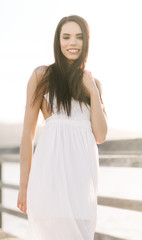 Fototapeta na wymiar Smiling young woman in a white dress standing on a wood pier during the golden hour sunset on a warm summer day.