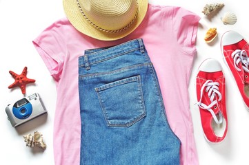 Flat lay women's travel outfit. Blue jeans, pink t-shirt, red gumshoes, sun hat, camera and seashells