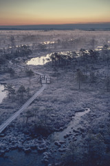 Obraz premium Wooden path, road in swamp on early winter morning with frozen trees and grass. Kemeri national park at sunrise with thick fog, Latvia. Vintage look.