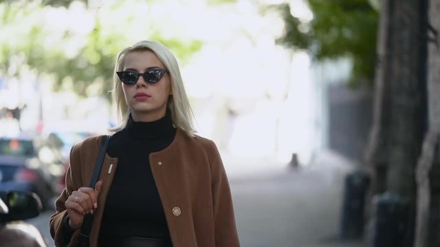 Portrait of young blonde businesswoman goes to work or date in autumn city. Girl have stylish look, sunglasses and nose piercing. Lady walking on the street alone. Fashion concept