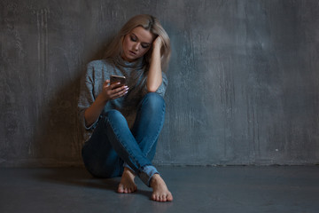 Helpline, psychological assistance. Suffering young woman sitting in a corner with a phone in her hand - obrazy, fototapety, plakaty