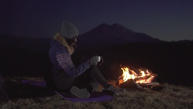 Camping young woman sitting and drinking from a Cup around the fire at night in the mountains,slow mo
