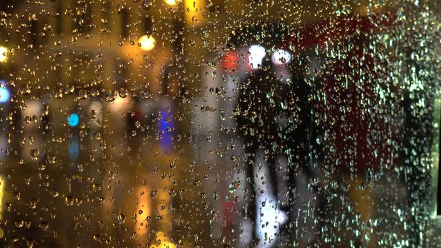 City street with people walking under the rain seen through a glass window with raindrops at night