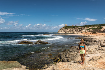 Attractive blond young woman in bathing suit on a rocky beach looking far away, blue sky and turquoise see in the background. Shot in Capo Mannu beach, Sardinia, Italy.